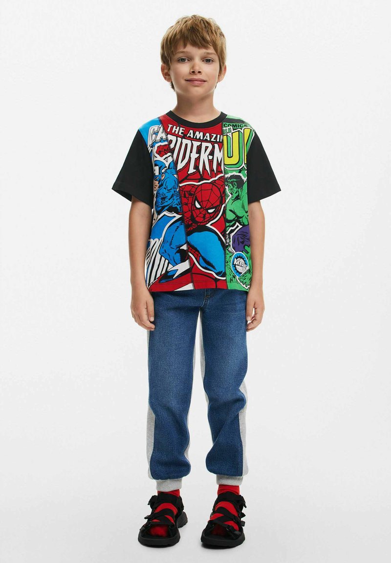 Short-sleeve multicolor T-shirt featuring comic book characters. Paired with blue jeans and black sandals with red accents.
