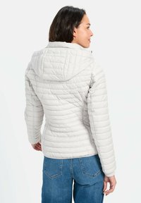Lys grå quiltet jakke med hætte, der har en tekstureret overflade og synlige syninger. Parret med blå jeans, der viser en afslappet pasform.