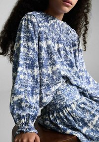 Blouse met blauw-witte bloemenprint, voorzien van een gathered halslijn en elastische taille. Lange pofmouwen, lichte stof en geruffelde manchetten.