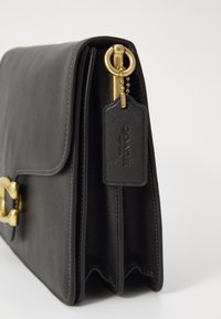 Sac à main en cuir noir avec une fermeture à rabat, quincaillerie dorée et une étiquette attachée par une chaîne. Texture lisse et forme structurée.