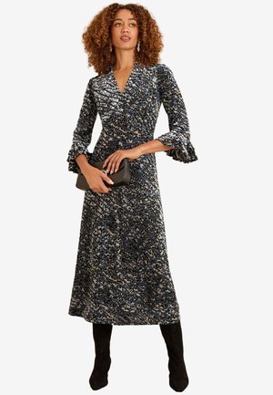 Donna con capelli ricci che indossa un vestito midi a maniche lunghe con fantasia, tiene una pochette nera e indossa stivali neri al ginocchio.