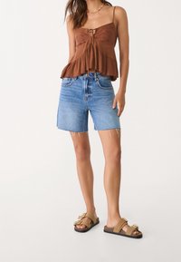 Ruskea, teksturoitu toppi, jossa on pitsikoristeita, yhdistetty vaaleansinisiin farkkushortseihin, joissa on rispaantuneet reunat. Jalkineina ovat vaaleanruskeat liukusandaalit.