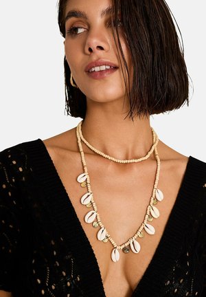 Femme aux cheveux courts mouillés portant un haut noir en tricot et plusieurs colliers superposés en perles beige, dont un avec des pendentifs en coquillage blanc et pièce d'or.