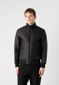 Melnā pūkainā jaka ar augstu apkakli, izšūtu tekstūru un priekšējo rāvējslēdzēju. Ribotās piedurknes un apakšmalā, piegriezta piegriezuma, viegla materiāla.