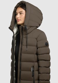 Cappotto imbottito marrone con ampio cappuccio, chiusura frontale con zip e tasche laterali. Presenta una texture trapuntata e un logo rotondo nero sulla manica.