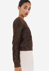 Cardigan marrone a maniche corte con una maglia texturizzata e scintillante. Presenta un collo rotondo e quattro bottoni sulla parte anteriore.