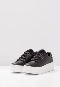 Černé kožené tenisky s tlustou bílou platformou, vyznačující se umělou kožešinou a černými tkaničkami s výrazem značky.