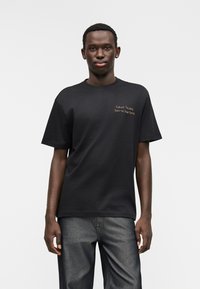 Μαύρο βαμβακερό μπλουζάκι με κοντά μανίκια και στρογγυλή λαιμόκοψη. Διαθέτει ανάγλυφη χρυσή κεντημένη φράση στο αριστερό στήθος.