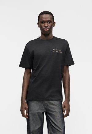 Black cotton t-shirt with short sleeves and a crew neckline. Features a textured gold embroidered phrase on the left chest.