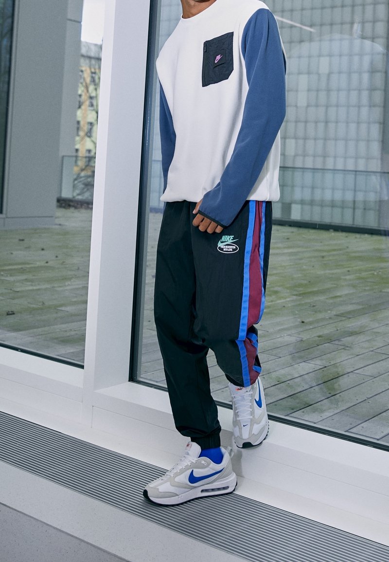 Vit sweatshirt med blå ärmar och ficka, svarta joggers med blå och marmor-ränder, samt vita sneakers med en blå swoosh.