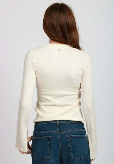 Top crema a maniche lunghe con texture morbida, design aderente e un piccolo logo vicino al collo, abbinato a jeans in denim blu scuro.