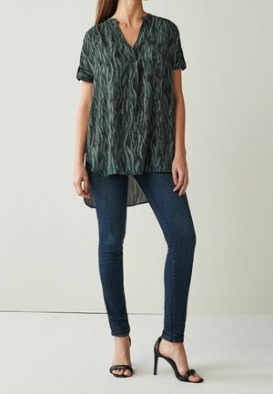Blouse à manches courtes, verte à motif, avec un col en V, une coupe fluide et un dos légèrement plus long. Associée à un jean skinny foncé et des talons ouverts.