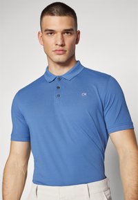 Camisola polo azul com uma frente de dois botões, mangas curtas e logótipo CK bordado no peito. Tecido liso e leve.