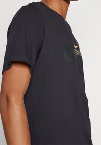 Černé bavlněné tričko s kulatým výstřihem, krátkými rukávy a jemným logem Nike v žluté barvě na přední straně. Hladká textura.