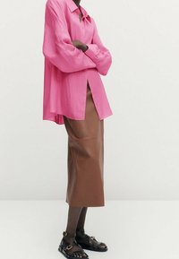Roze oversized shirt met een puntige kraag, gecombineerd met een bruin leren midi-rok en zwarte sandalen met banden, versierd met gouden accenten.