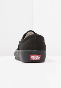 Zapatilla de lona negra con punta redonda, suela plana y acentos de goma. Presenta una plantilla blanca y un logo rojo en el talón.