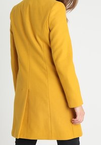 Geel wolmix jas met lange mouwen, slim fit, rugsplit en minimale stikseldetails, gezien vanaf de achterkant.
