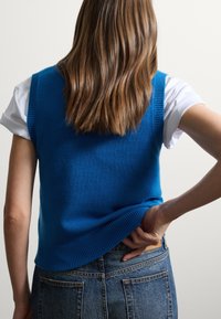 Gilet di maglia blu con orlo a costine, indossato sopra una camicia bianca. La vista posteriore mostra la texture e la silhouette, abbinata a jeans a vita alta.