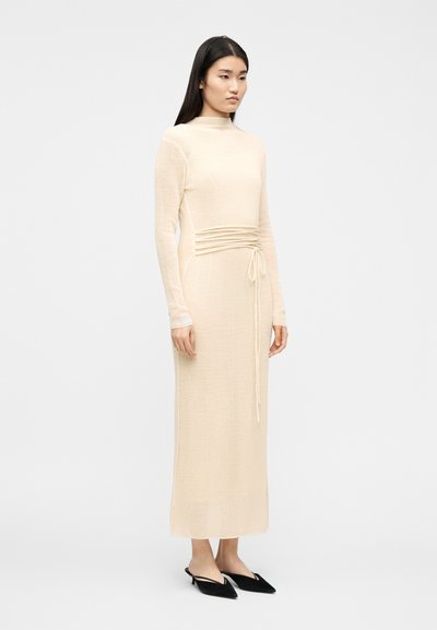Femme debout portant une robe en maille beige à manches longues avec un détail de lien à la taille et des mules à talons noires à bouts pointus.