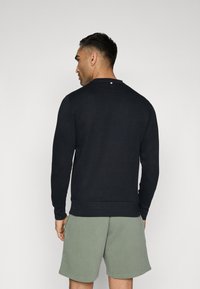 Mornarska crewneck dukserica iz mehkega blaga, z ribastimi manšetami in robom, s majhnim poudarkom na zadnjem delu vratu. Obuta z zelenimi kratkimi hlačami.