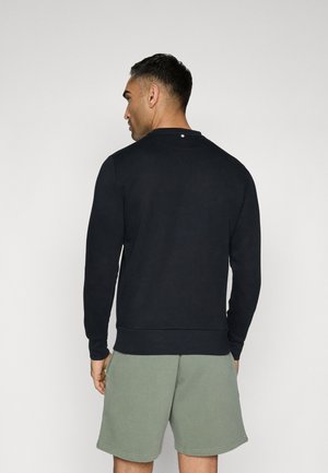Mand iført en sort sweatshirt med lange ærmer og lysegrønne shorts, set bagfra mod en ensartet lys baggrund.