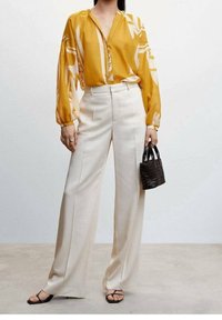 Gelbe Bluse mit auffälligem weißen Blumenmuster, langen Ärmeln und V-Ausschnitt. Weiße weit geschnittene Hosen. Frau hält eine kleine schwarze geflochtene Handtasche.