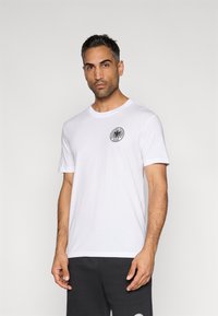 Λευκό κοντομάνικο μπλουζάκι κατασκευασμένο από βαμβάκι, με ένα κυκλικό μαύρο λογότυπο στο αριστερό στήθος. Στρογγυλή λαιμόκοψη και απλός σχεδιασμός.