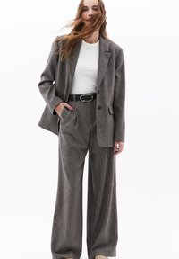 Harmaa räätälöity puku, jossa on hieman ylisuuri bleiseri ja leveät housut, joissa on teksturoitu kangas sekä vyö kultaisella soljella.