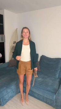 Tumši zaļš kardigans pār krēmkrāsas topu, kombinēts ar bēša krāsas šortiem. Apavi ir gaiši bēšas krāsas augstpapēžu kurpes. Fonā redzams zilas krāsas dīvāns un sērfošanas dēlis.