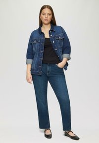 Veste en denim bleu foncé avec des manches repliées bleu clair, associée à un haut noir et un jean droit bleu foncé. Ballerines noires aux pieds.
