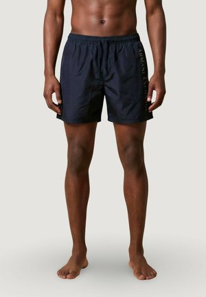 Hombre con bañador negro de Armani Exchange, de pie descalzo contra un fondo liso de color gris claro.