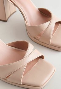 Sandales à talons beiges à bouts ouverts avec larges brides croisées et devant carré, présentées sur une surface claire.