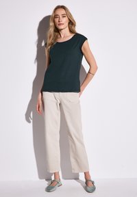 Tmavě zelený top s krátkým rukávem spárovaný se světle béžovými kalhotami rovného střihu a tyrkysovými plochými botami, vyrobený z hladké látky a s volným střihem.