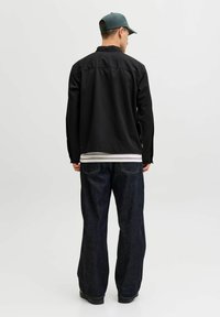 Uomo con indosso una maglia nera a maniche lunghe, jeans larghi a gamba larga di colore scuro, camicia a righe bianca sotto e cappellino verde, in piedi di spalle su sfondo bianco.