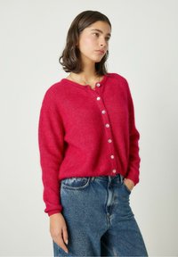 Cárdigan rosa difuso con cuello redondo, cierre de botones y puños acanalados, combinado con unos vaqueros de mezclilla azul. Textura suave y color vibrante.