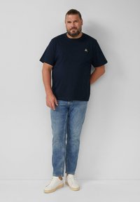 Man met een baard, gekleed in een marineblauw t-shirt, lichtblauwe jeans en witte sneakers, staande tegen een effen lichtgrijze muur.