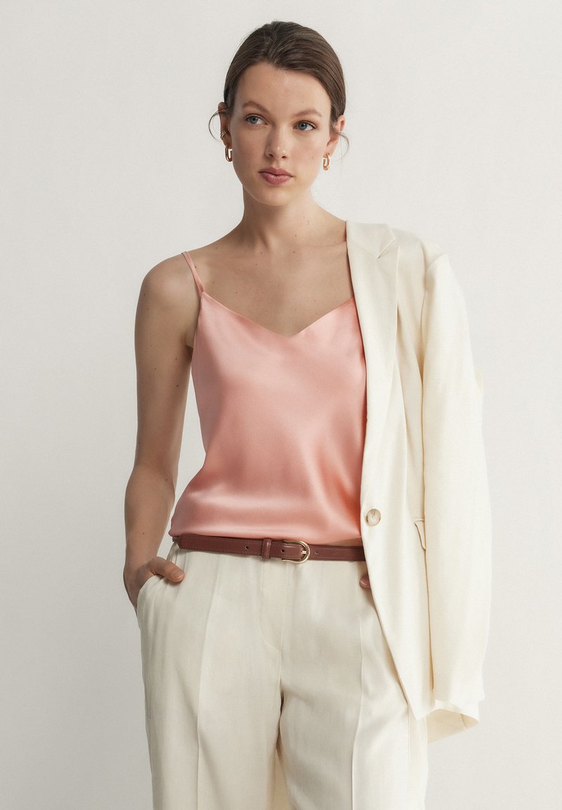 Femme portant un caraco en satin rose, un pantalon taille haute crème avec une ceinture marron, et un blazer crème posé sur son épaule.