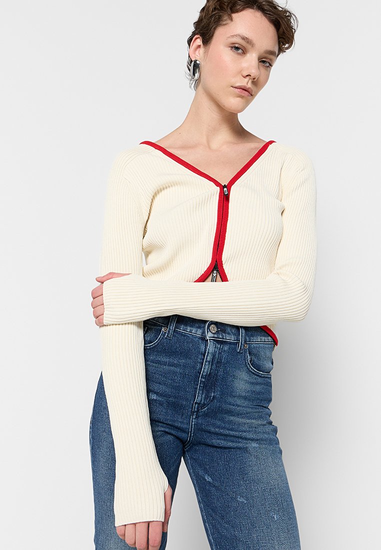Cardigan crema a coste con zip e rifinitura rossa; maniche lunghe con fori per il pollice; abbinato a jeans in denim di lavaggio medio.