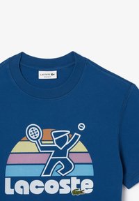 Camiseta de algodón azul con un gráfico de un jugador de tenis sobre un fondo de rayas multicolores. Logo de Lacoste en la parte inferior.