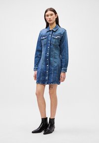 Denim φόρεμα σε σκούρο μπλε με μακριά μανίκια, κλείσιμο μπροστά με κουμπιά, δύο τσέπες στο στήθος και φθαρμένο στρίφωμα. Φοριέται με μαύρες μπότες μέχρι τον αστράγαλο.