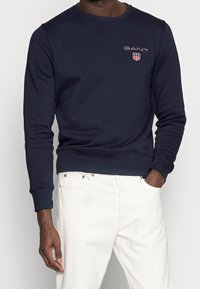 Donkerblauwe crewneck-trui gemaakt van zacht katoen, voorzien van een klein GANT-logo en schildgrafiek op de borst. Geribbelde manchetten en zoom.