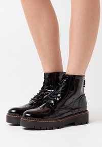 Black patent leather ankle boots with a brogue design, featuring a chunky platform sole and lace-up front. Textured finish and detailed stitching.