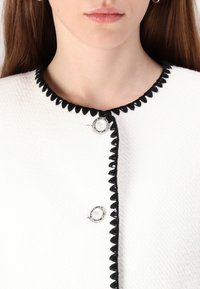 Kvinna iklädd en vit texturerad cardigan med svarta vågiga detaljer och två dekorativa pärlknappar nära halsringningen.