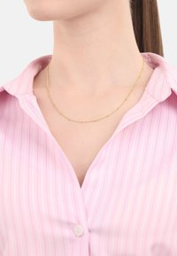 Hängsmycke i guldton med en delikat design och små, jämnt fördelade pärlor. Bärs över en ljusrosa randig skjorta.