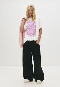 En blond kvinde holder en lyserød jakke over skulderen, iført en hvid t-shirt med teksten "Endless Coast," brede sorte bukser, hvide sneakers og en sølvfarvet bæltespænde.