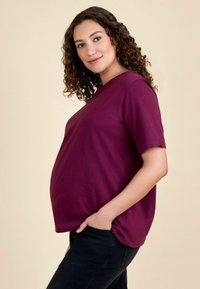 Bordeaux zwangerschap t-shirt met korte mouwen, ontspannen pasvorm, effen kleur en een gladde textuur; gecombineerd met donkere jeans tegen een neutrale achtergrond.