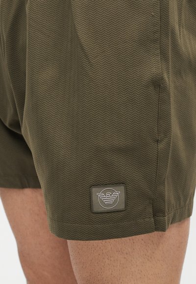 Κοντινό πλάνο λαδί σορτς με διακριτικό σχέδιο chevron και μικρό ορθογώνιο λογότυπο κοντά στο στρίφωμα στο πάνω μέρος του μηρού ενός ατόμου.