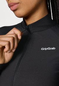 Juoda dviratininko striukė su aukštu apykakle, priekiniais užtrauktukais ir balta "GripGrab" logotipu. Lygus, elastingas audinys, glaudus pjūvis ir jokių matomų raštų.