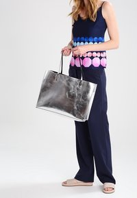 Stříbrná kovová taška přes rameno se strukturovaným tvarem, ramenními popruhy a hladkým povrchem. Nositá s námořnickým topem a tmavými kalhotami.
