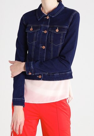 Persoon draagt een donkerblauwe spijkerjas over een lichtroze blouse en felrode broek, met één arm gekruist over de romp.